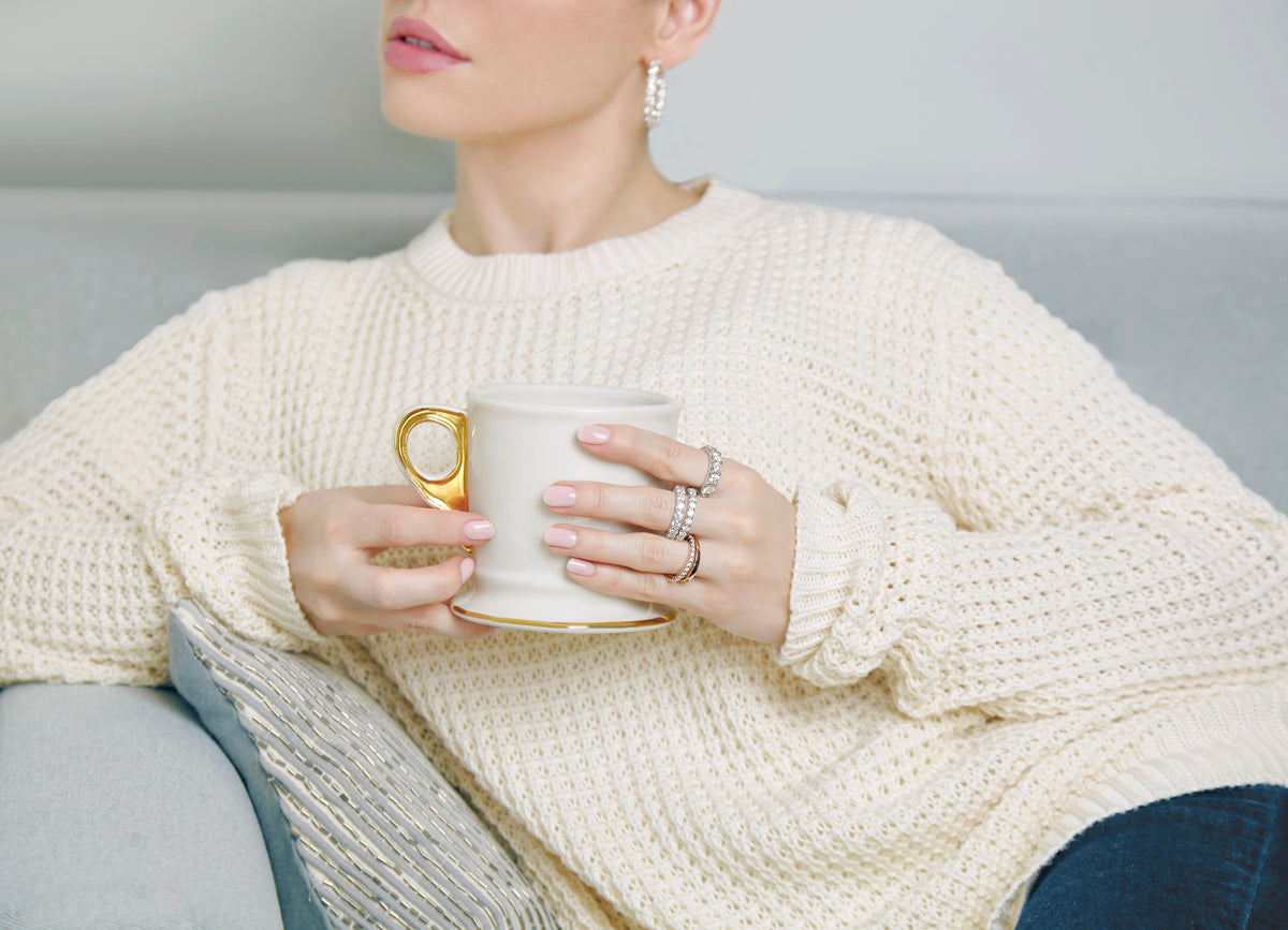 Hands holding a white mug, wearing stacked diamond rings and a gold ring